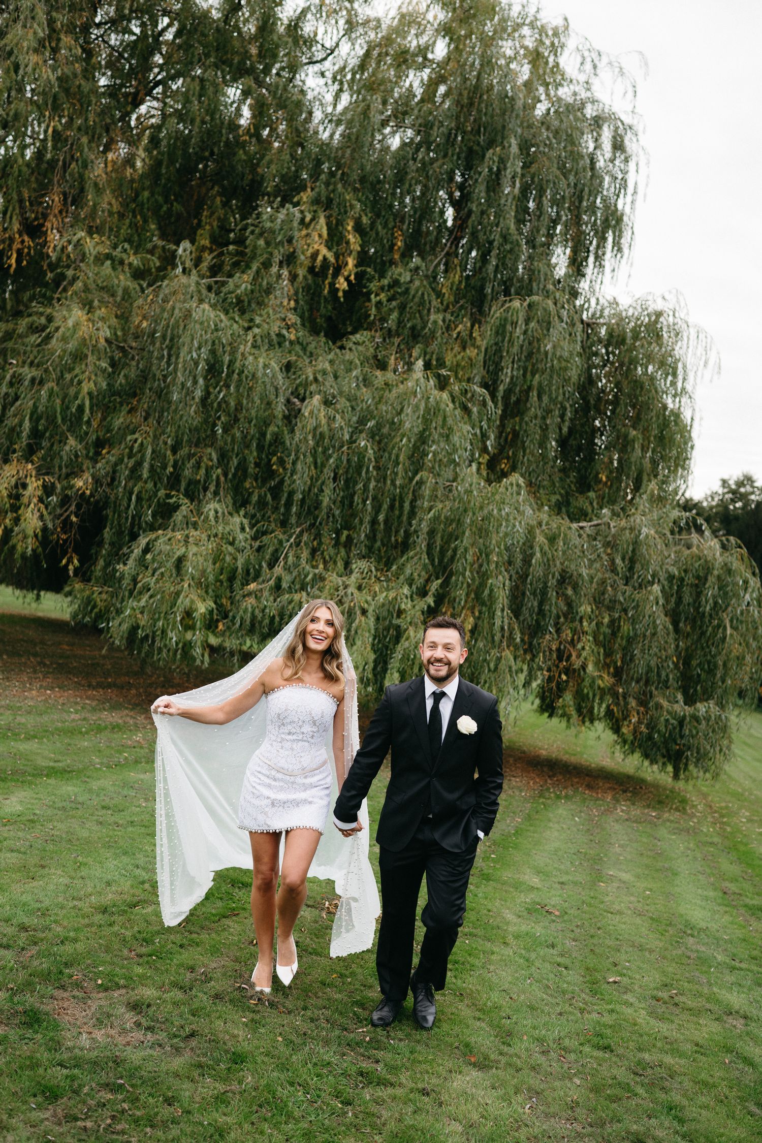 A couple poses under a large weeping willow tree on a grassy lawn during their wedding celebration.