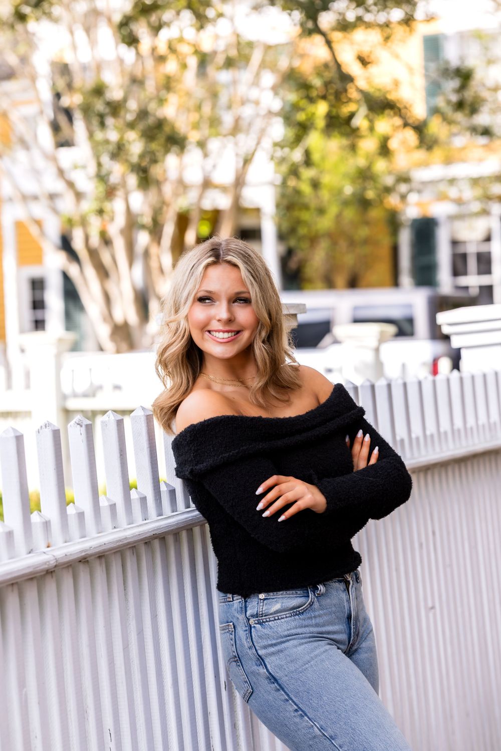 A person in a black off-shoulder top and jeans poses casually against a white picket fence on a sunny day.