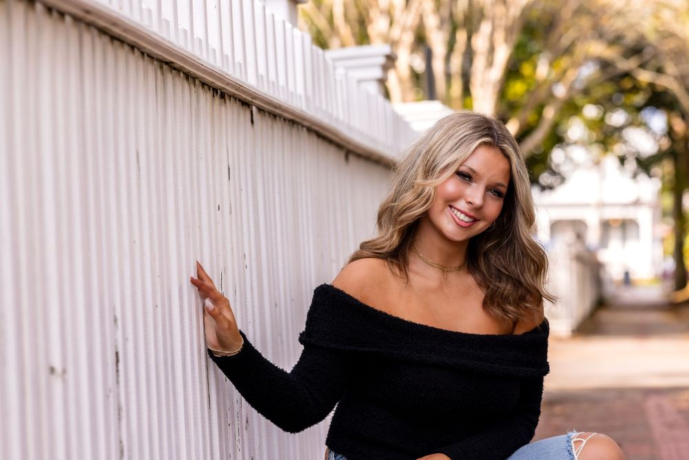 Woman in black off-shoulder top poses against white wall in outdoor photoshoot series.
