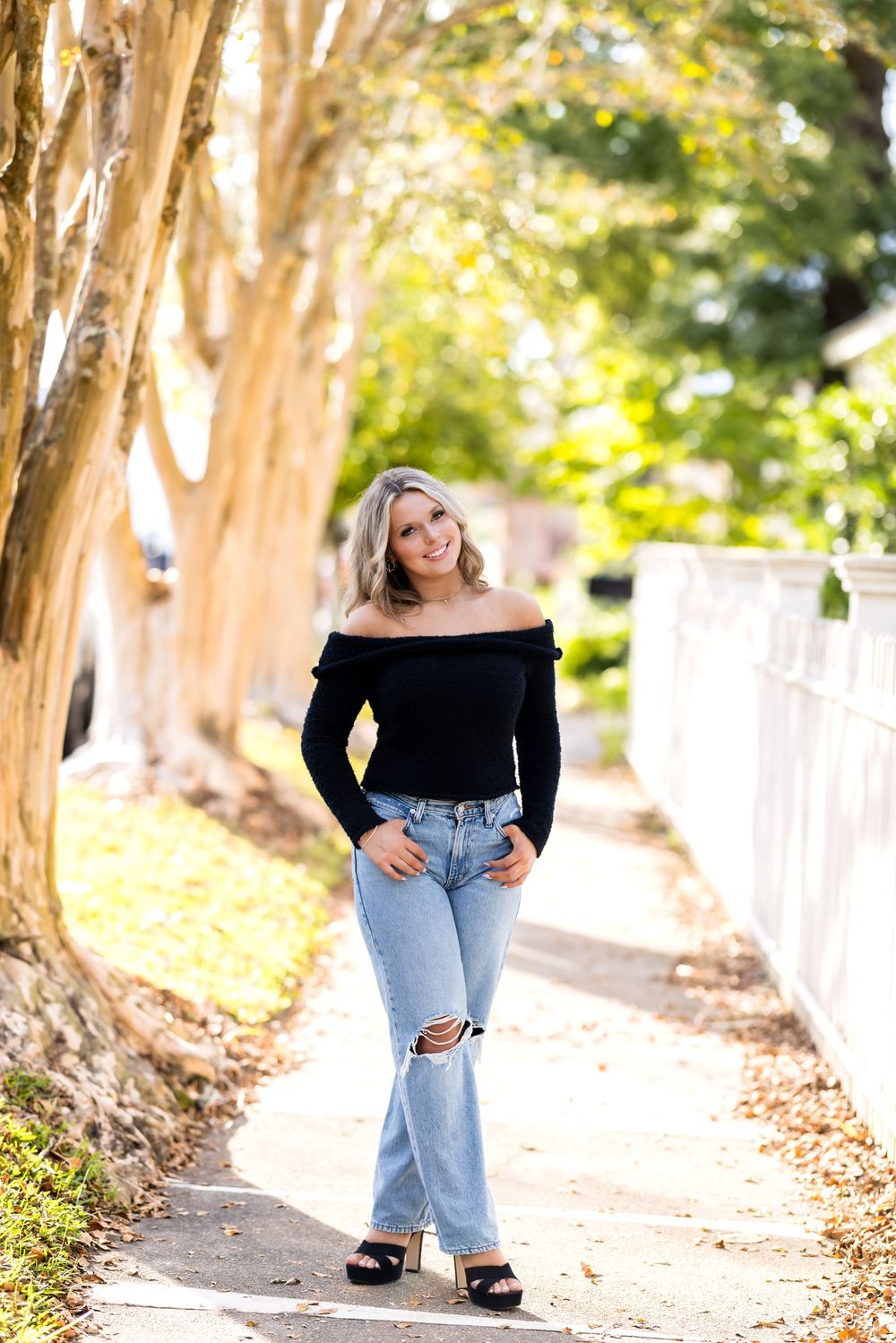 A woman in ripped jeans and black top poses along a tree-lined path in autumn sunlight.
