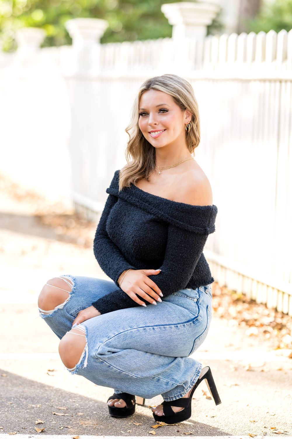 A woman in a black off-shoulder top and ripped jeans poses outdoors in a casual crouching position.