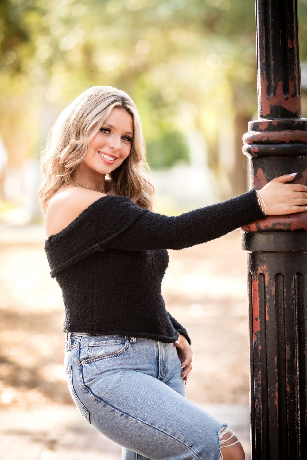 A person in a black sweater and jeans poses playfully against a dark pole outdoors in warm lighting.