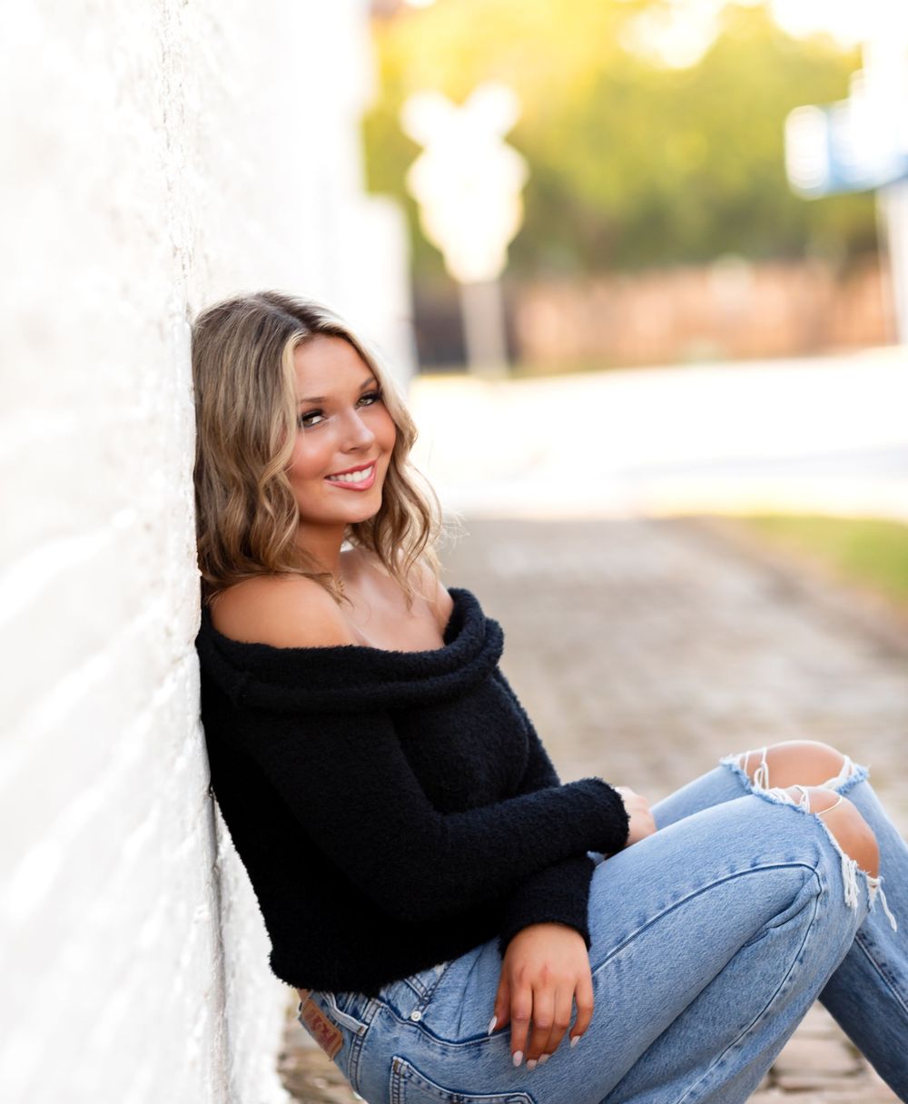 Woman in black off-shoulder sweater and ripped jeans poses casually outdoors during golden hour.