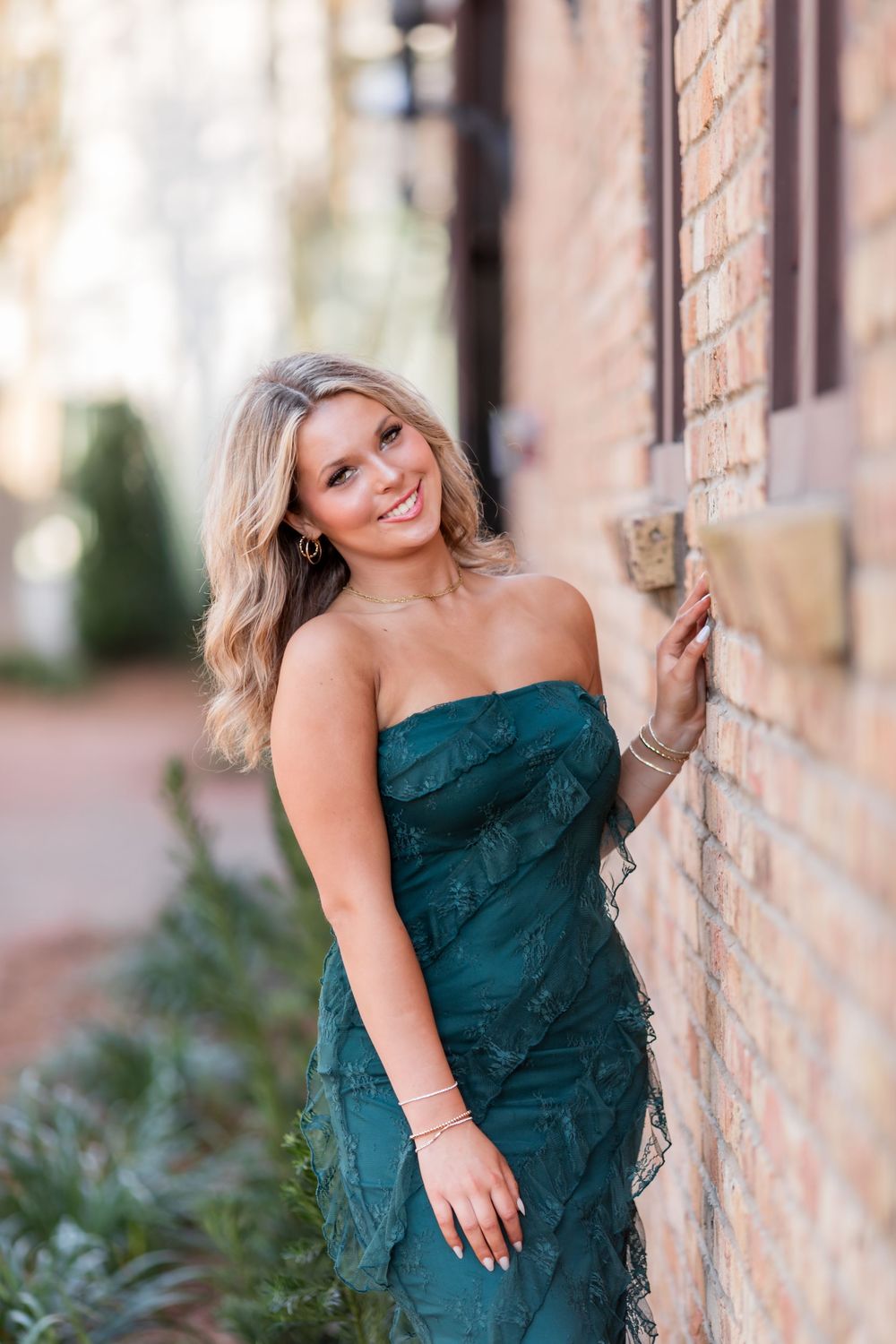 A woman in an elegant dark green formal dress poses against a brick wall in a series of outdoor portraits.
