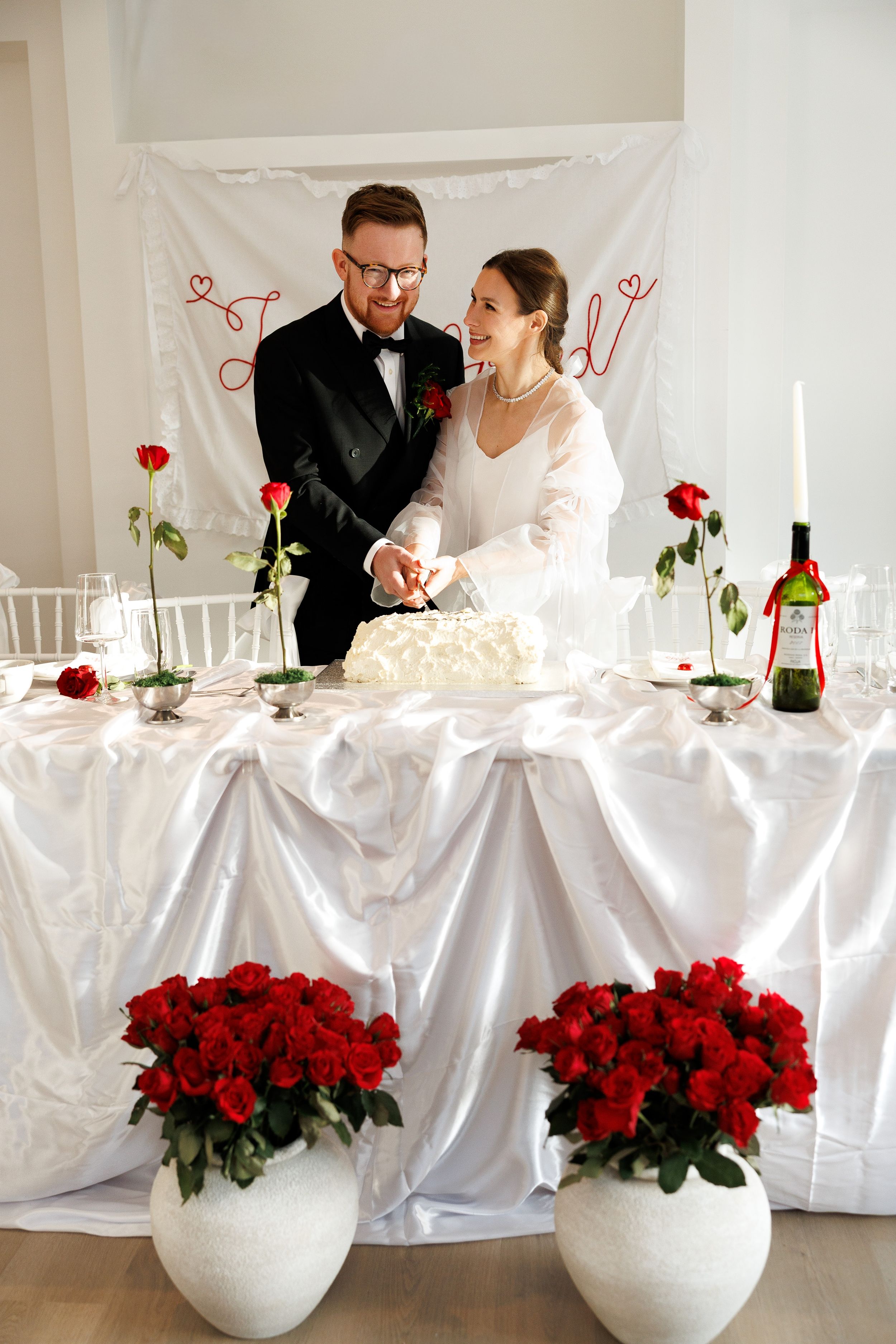 Bride and groom cutting white wedding cake at decorated table with red roses and candles.