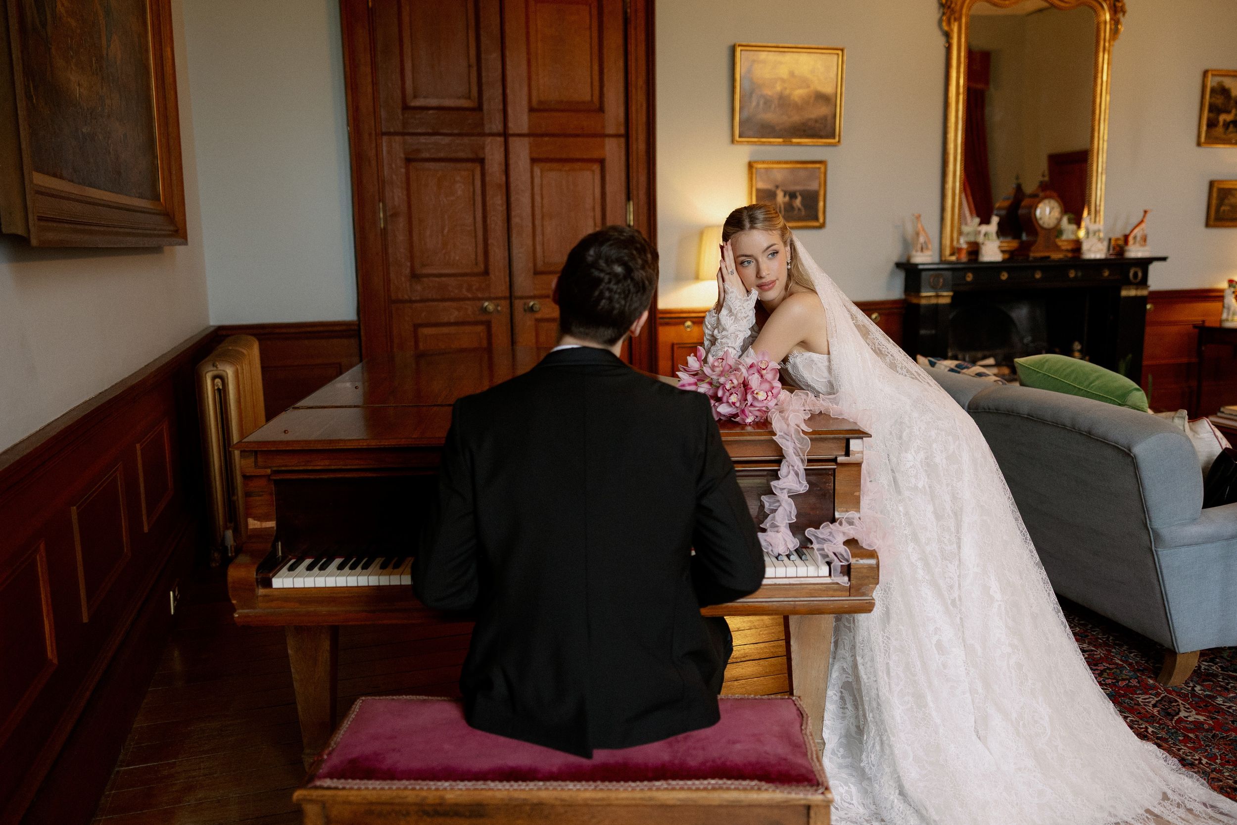 Bride in white gown leans on piano as groom plays during elegant wedding reception in classic interior.