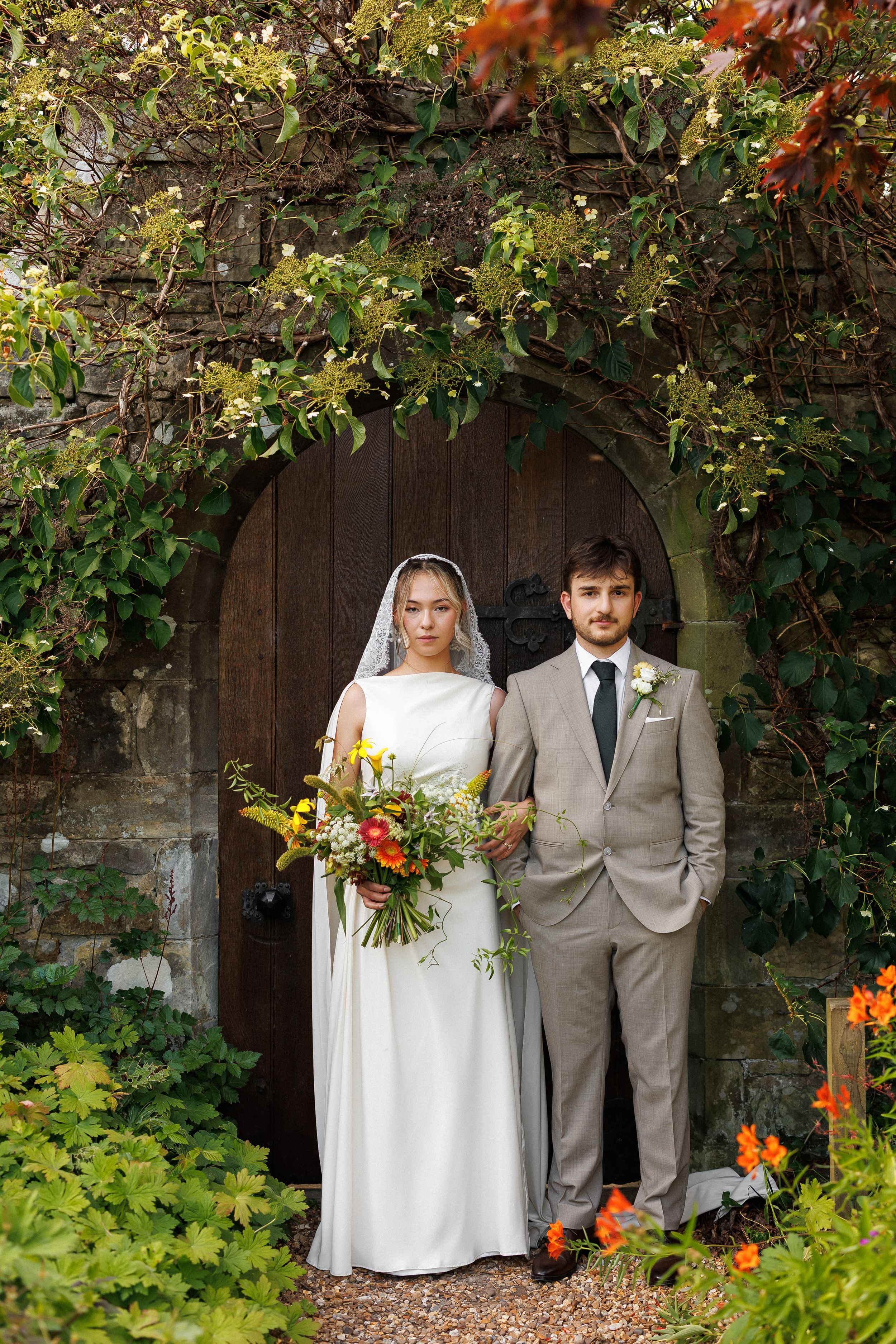 Bride and groom stand before ivy-covered stone arch with wooden door, she holds colorful bouquet, he wears gray suit.