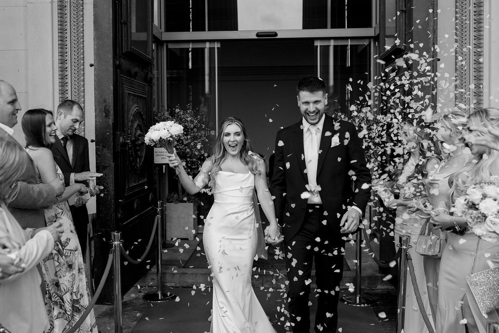 A newlywed couple exits their wedding ceremony while guests throw white confetti in celebration.