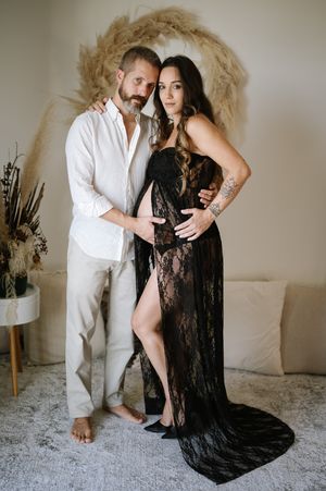A romantic maternity photoshoot featuring a couple posing against a beige backdrop with a circular dried palm leaf wreath.