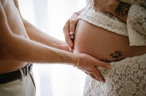 Close-up shot of hands gently resting on a pregnant belly with delicate lace details and visible tattoos.