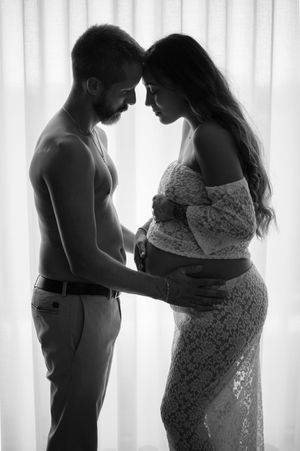 Intimate black and white maternity photo of a couple in tender embrace by a window with soft backlighting.