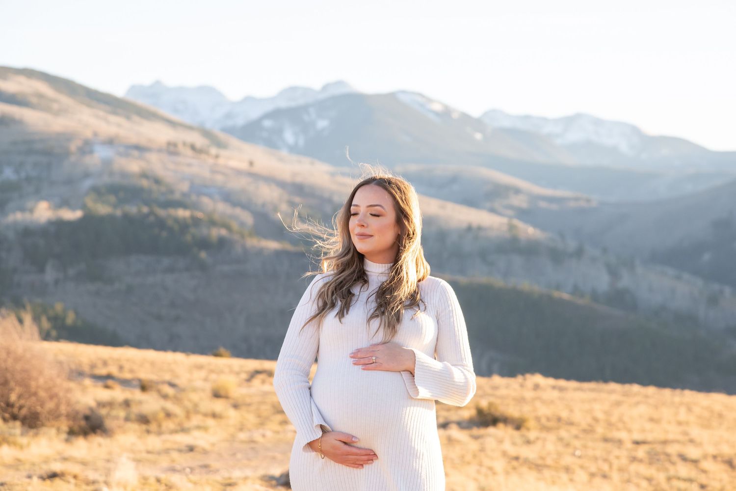 Poetic and beautiful mother-to-be closes her eyes and holds her pregnant belly during golden hour with mountain peaks in the background Vail