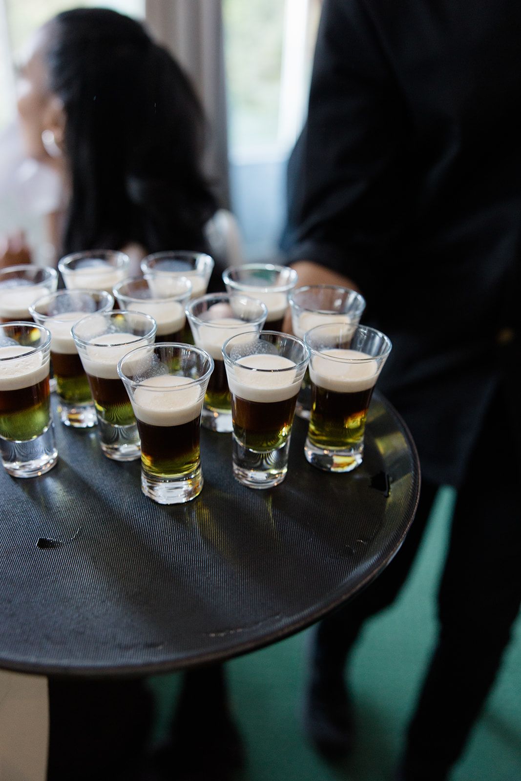 A black serving tray holds multiple small beer tasting glasses filled with dark and light craft beers with foamy heads.
