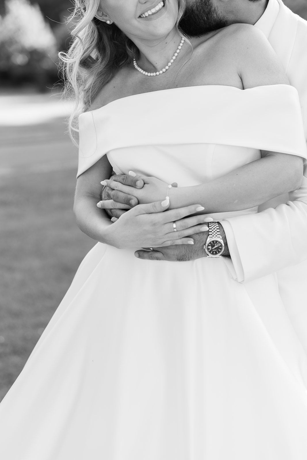 A couple in wedding attire embrace closely, with the bride wearing a white gown and pearl necklace in an outdoor setting.