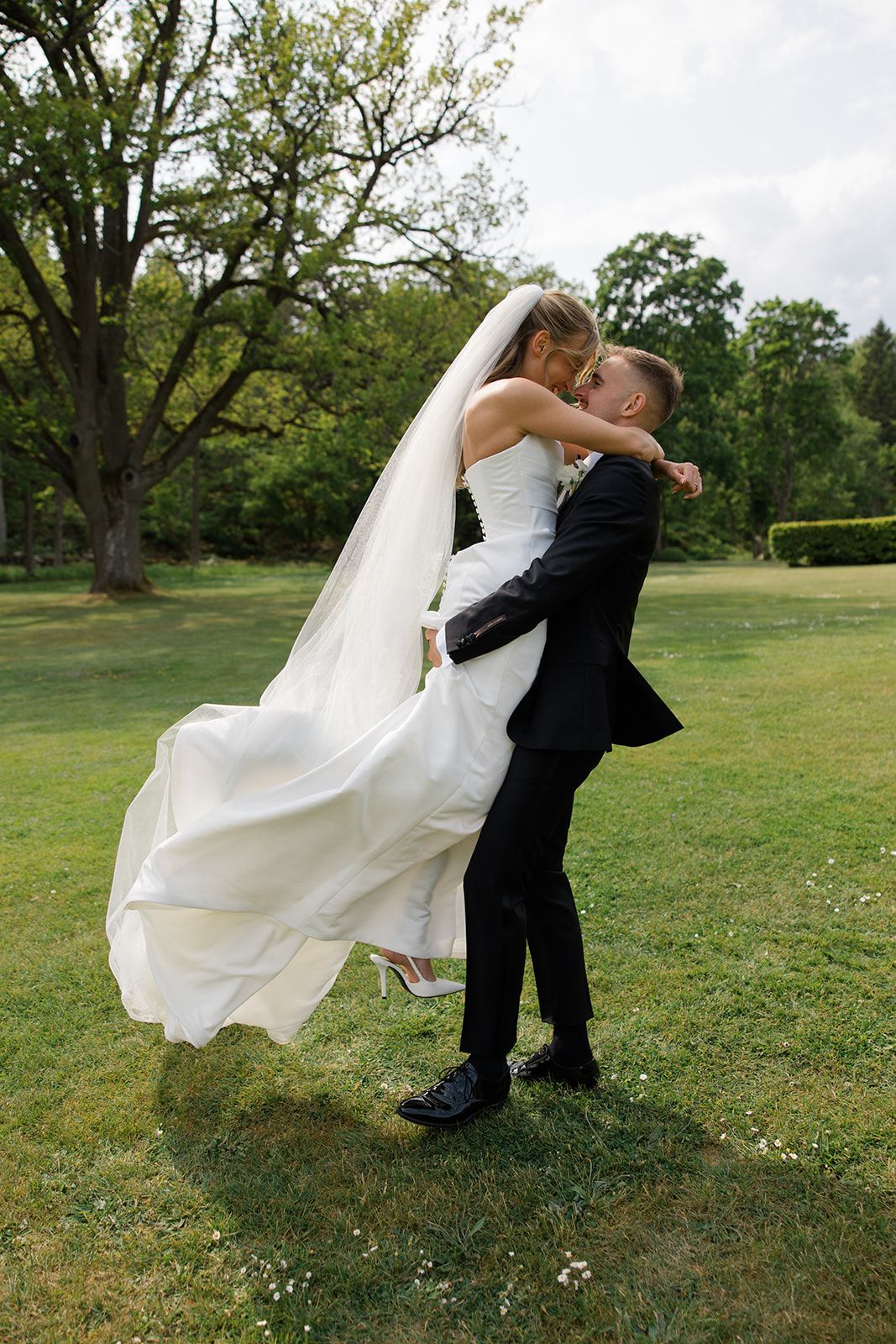 A couple embraces on their wedding day outdoors on a grassy lawn with trees in the background.