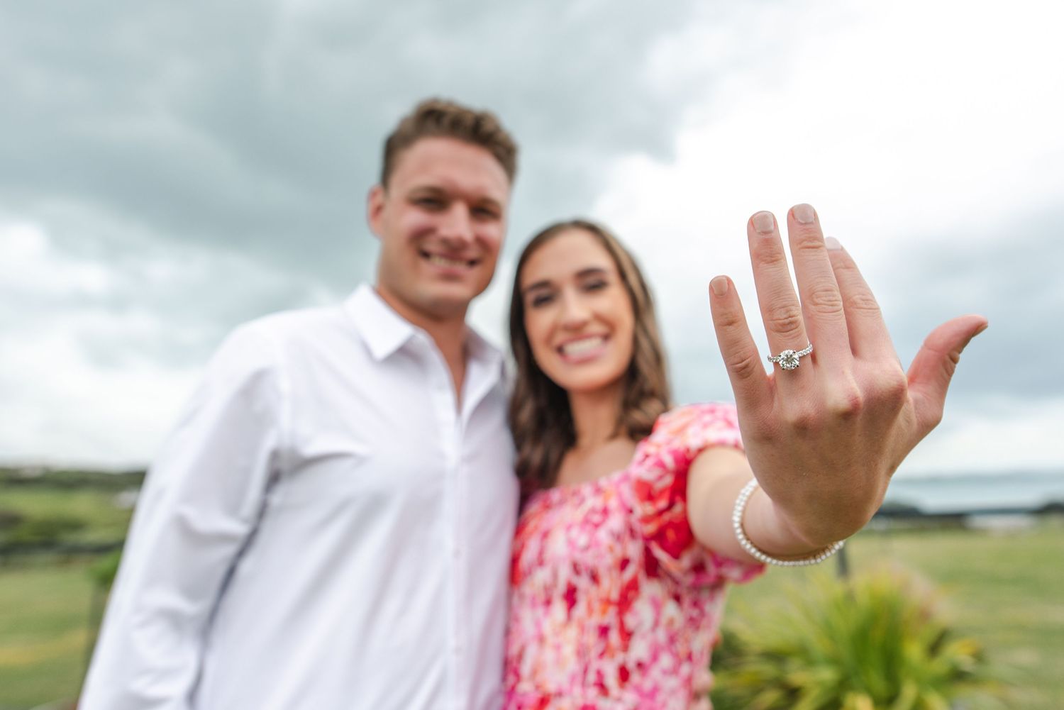 A couple in white and pink attire poses joyfully in a green outdoor setting on a cloudy day.