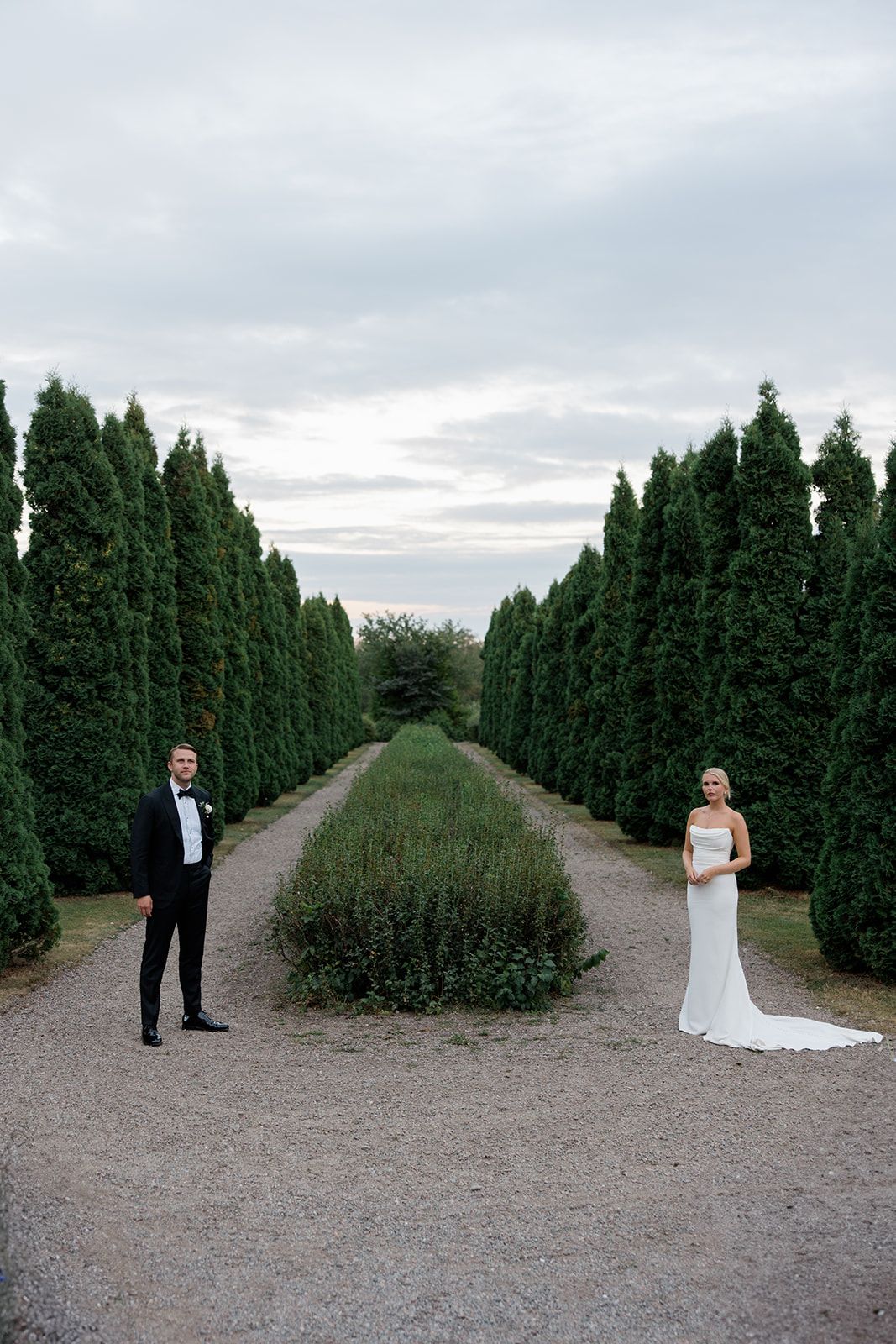A couple stands apart on a gravel path lined with tall evergreen hedges under an overcast sky.