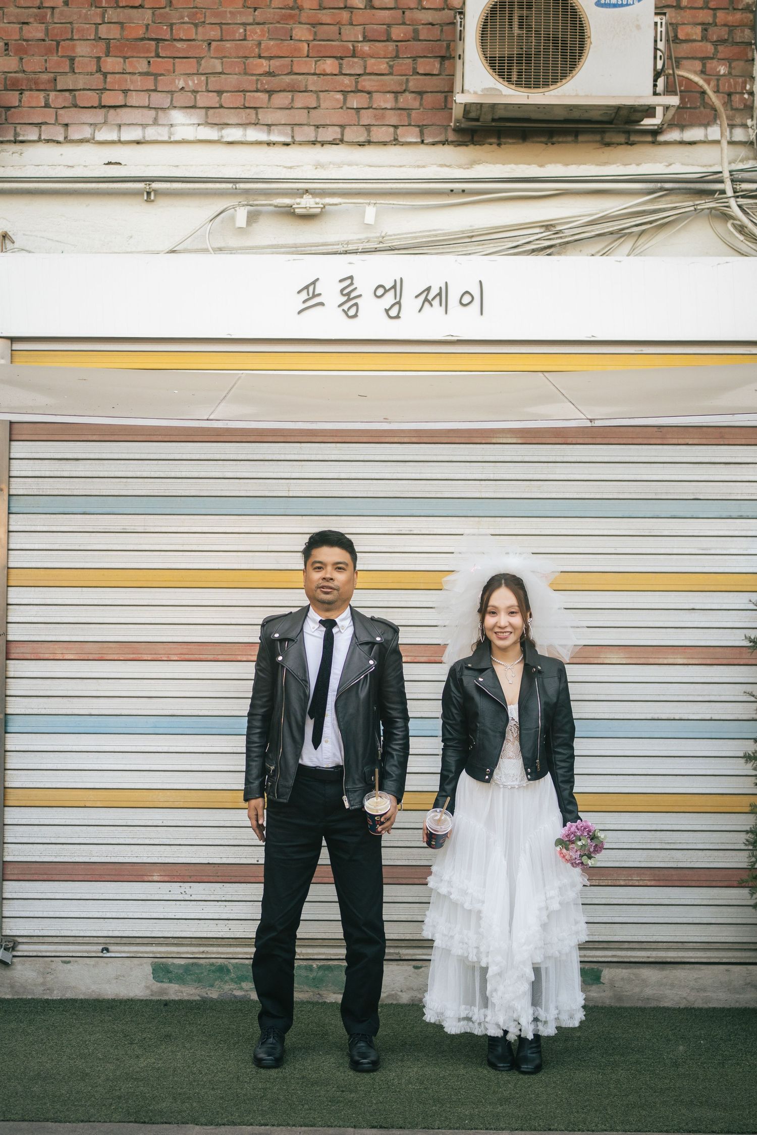 A couple in contrasting formal wear stands against a metal shutter wall with Korean text and an AC unit above.