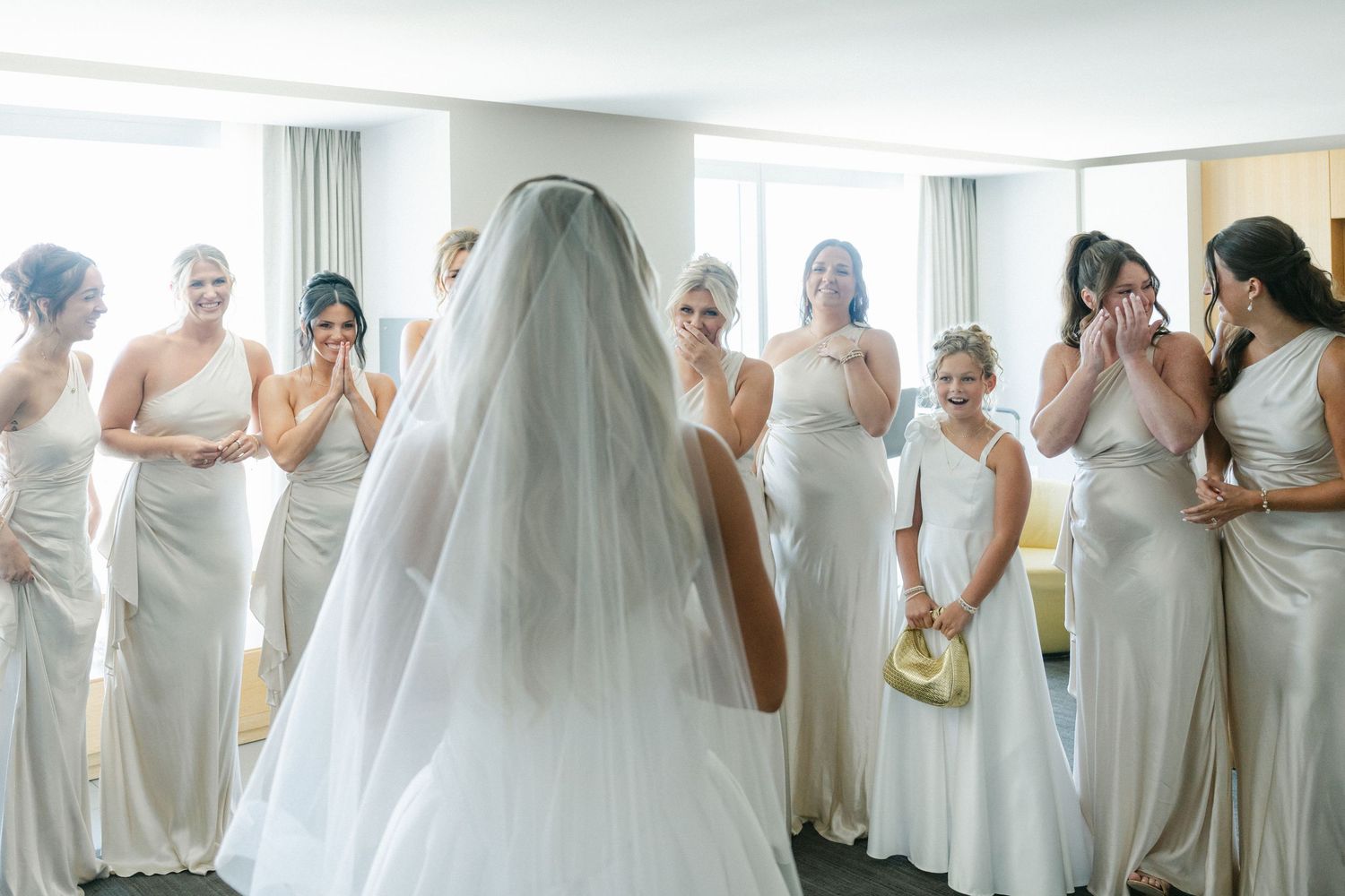 A group of bridesmaids in white dresses gather around a bride in a bright room during wedding preparations.