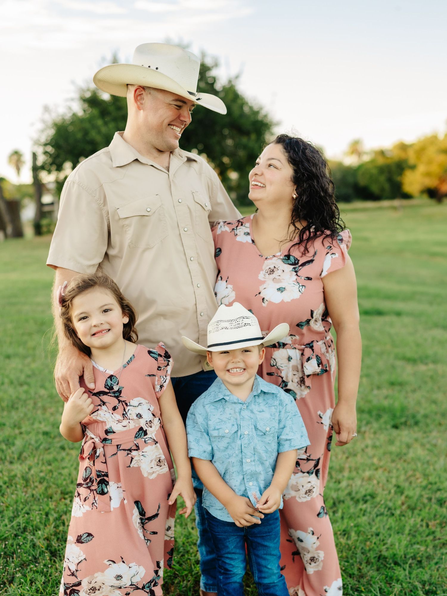 A family wearing coordinated outfits of pink floral dresses and cowboy attire poses together in a grassy field at sunset.