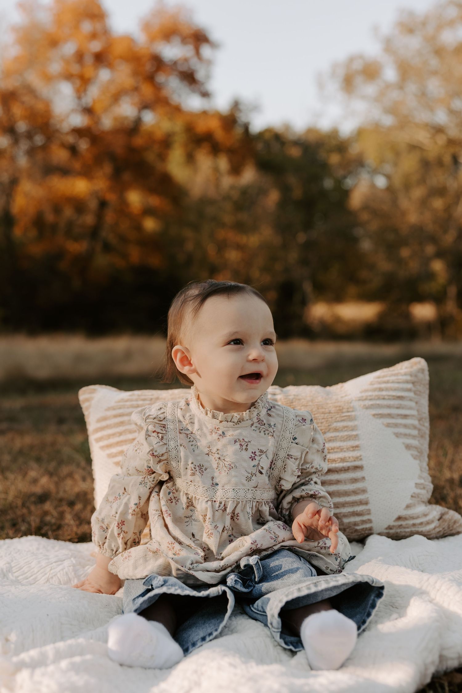Baby in lace outfit sitting on white blanket during autumn outdoor photo session.