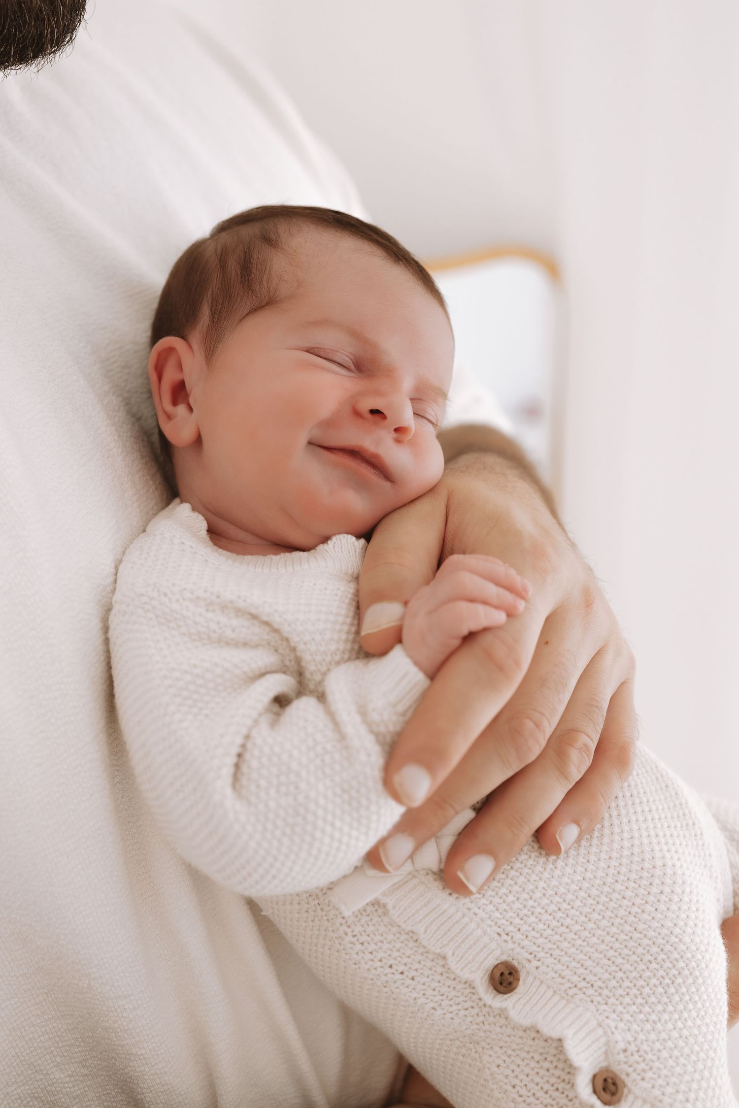 A sleeping newborn baby swaddled in white knit fabric with wooden buttons, photographed in a minimalist style.