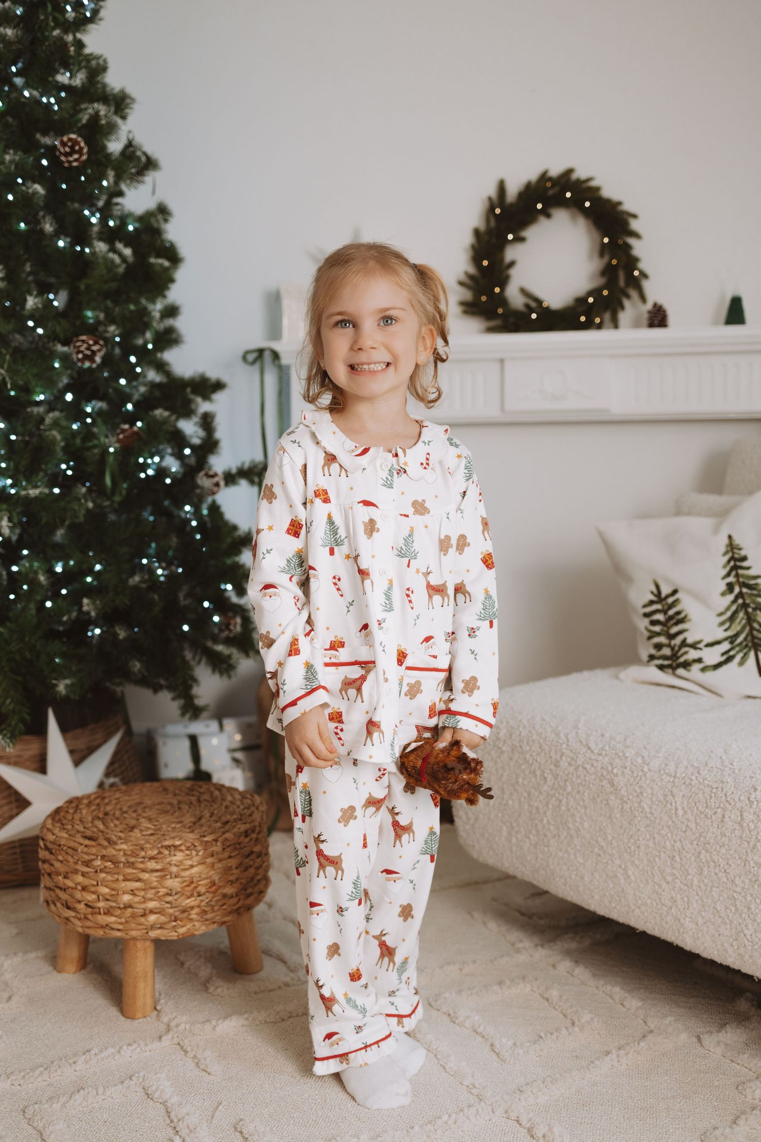 Festive holiday scene with child in white reindeer print pajamas sitting by decorated Christmas tree and wreath decor.