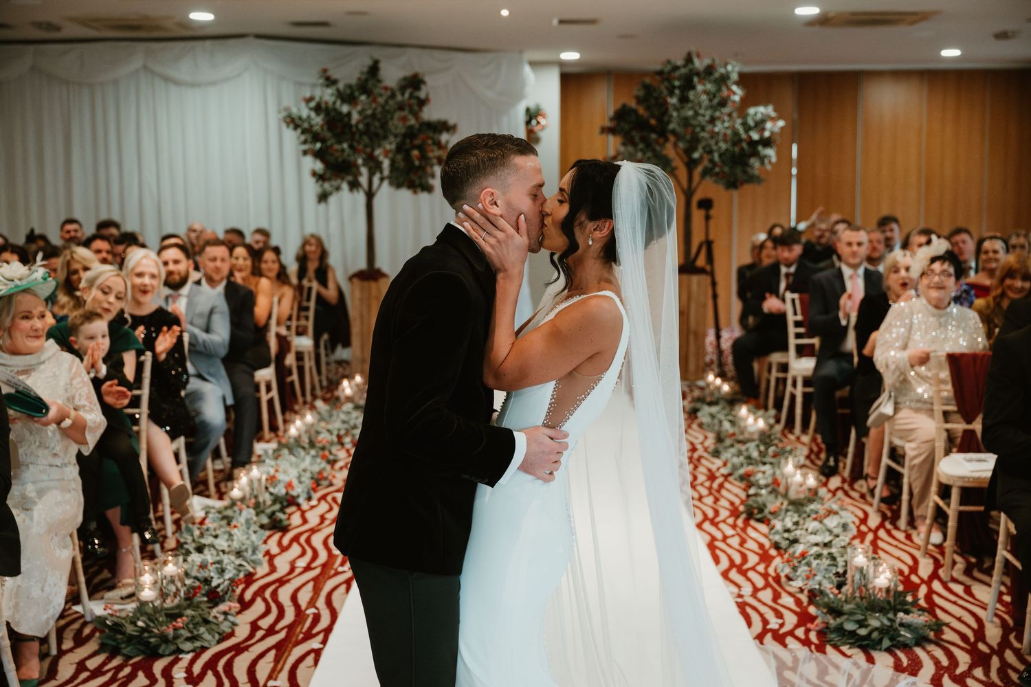 A first kiss during an indoor wedding ceremony with floral aisle decor and guests watching at bagden hall. photographer Daniel Berry