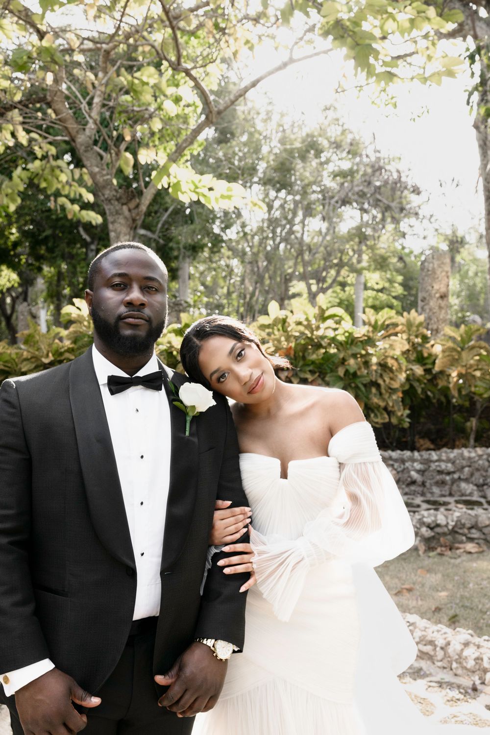 A romantic wedding couple shares an intimate moment outdoors against a backdrop of trees and stone walls.