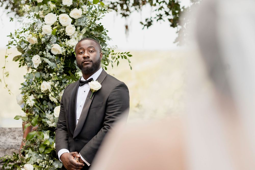 A person in a black suit poses elegantly against a backdrop of white floral arrangements and greenery.