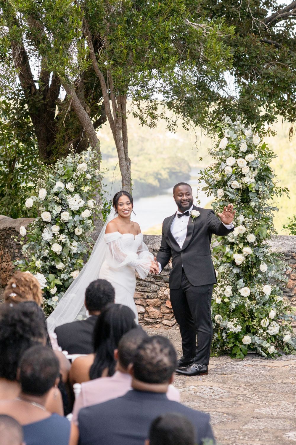 Newlyweds celebrate during an outdoor wedding ceremony surrounded by trees and white floral arrangements.