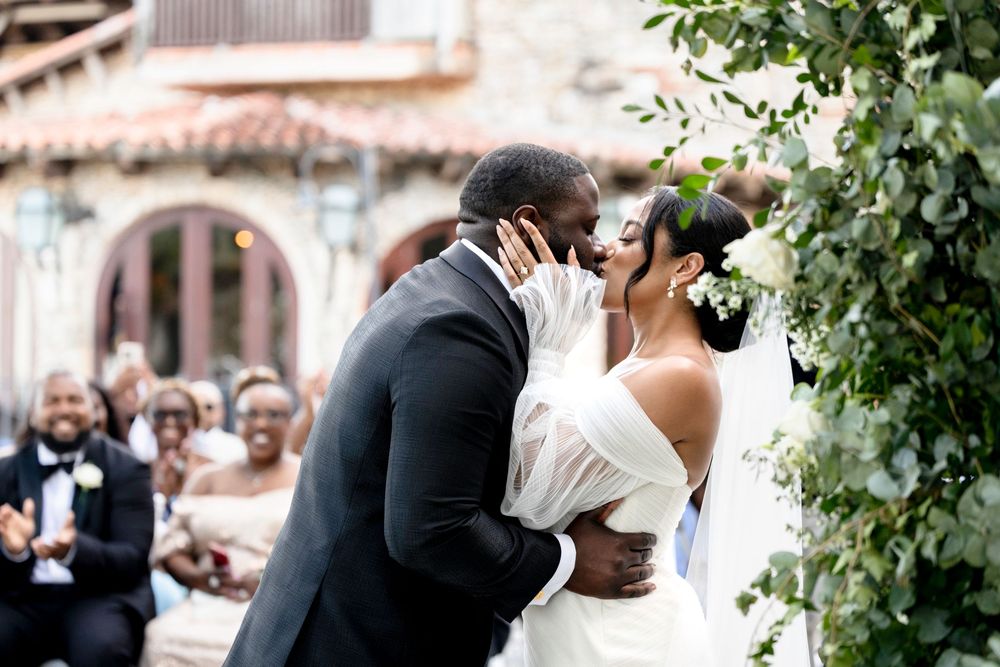 A romantic wedding kiss moment outdoors with greenery and architecture in the background.