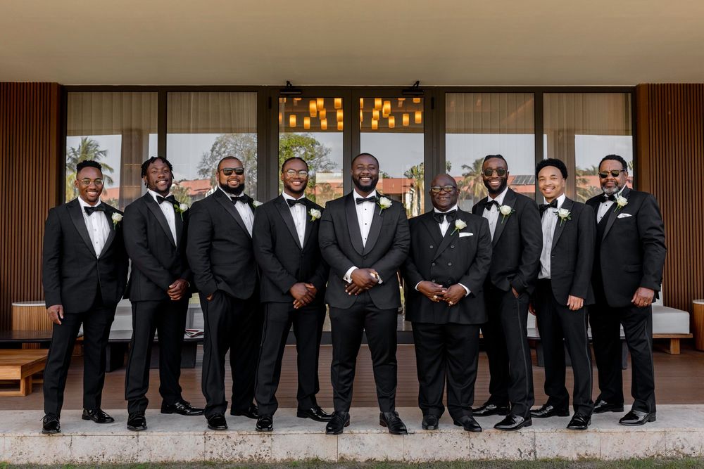 A group in formal black tuxedos and bow ties stand together in a row for a wedding party photo.