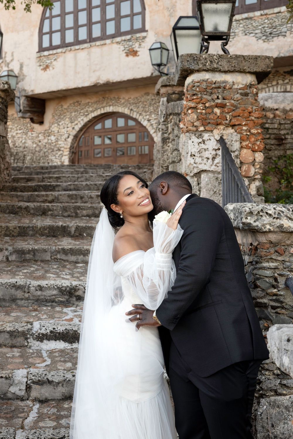 Romantic wedding couple embraces against a rustic stone wall backdrop with arched windows.