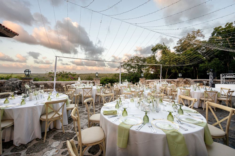 Elegant outdoor wedding reception setup with white tablecloths and green accents under a white tent at sunset.