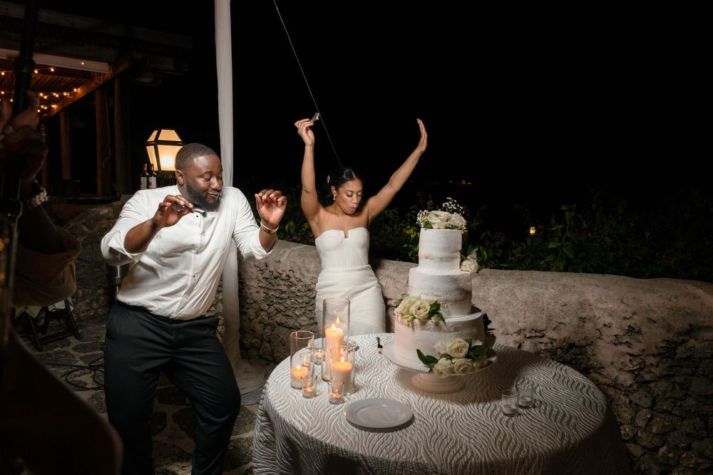 Couple celebrates energetically next to their elegant white wedding cake at an outdoor evening reception.
