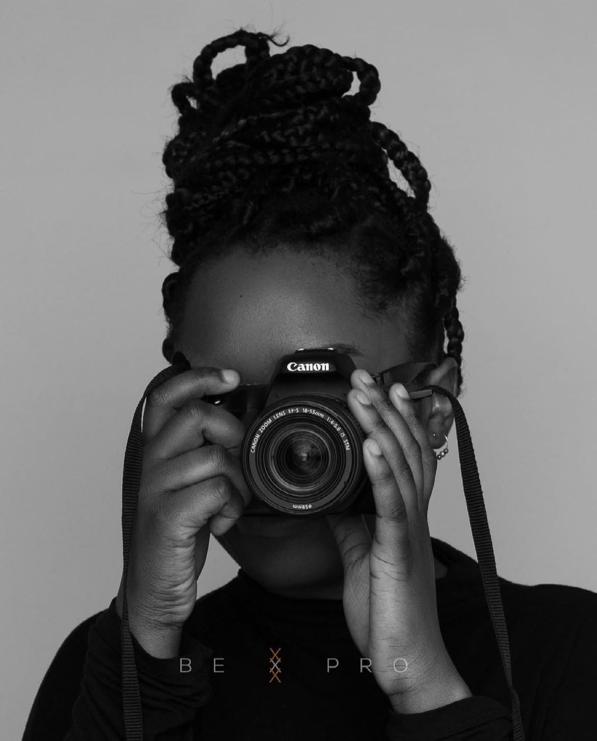 Black and white portrait of a Black woman with braided updo holding a Canon DSLR camera up to her face.