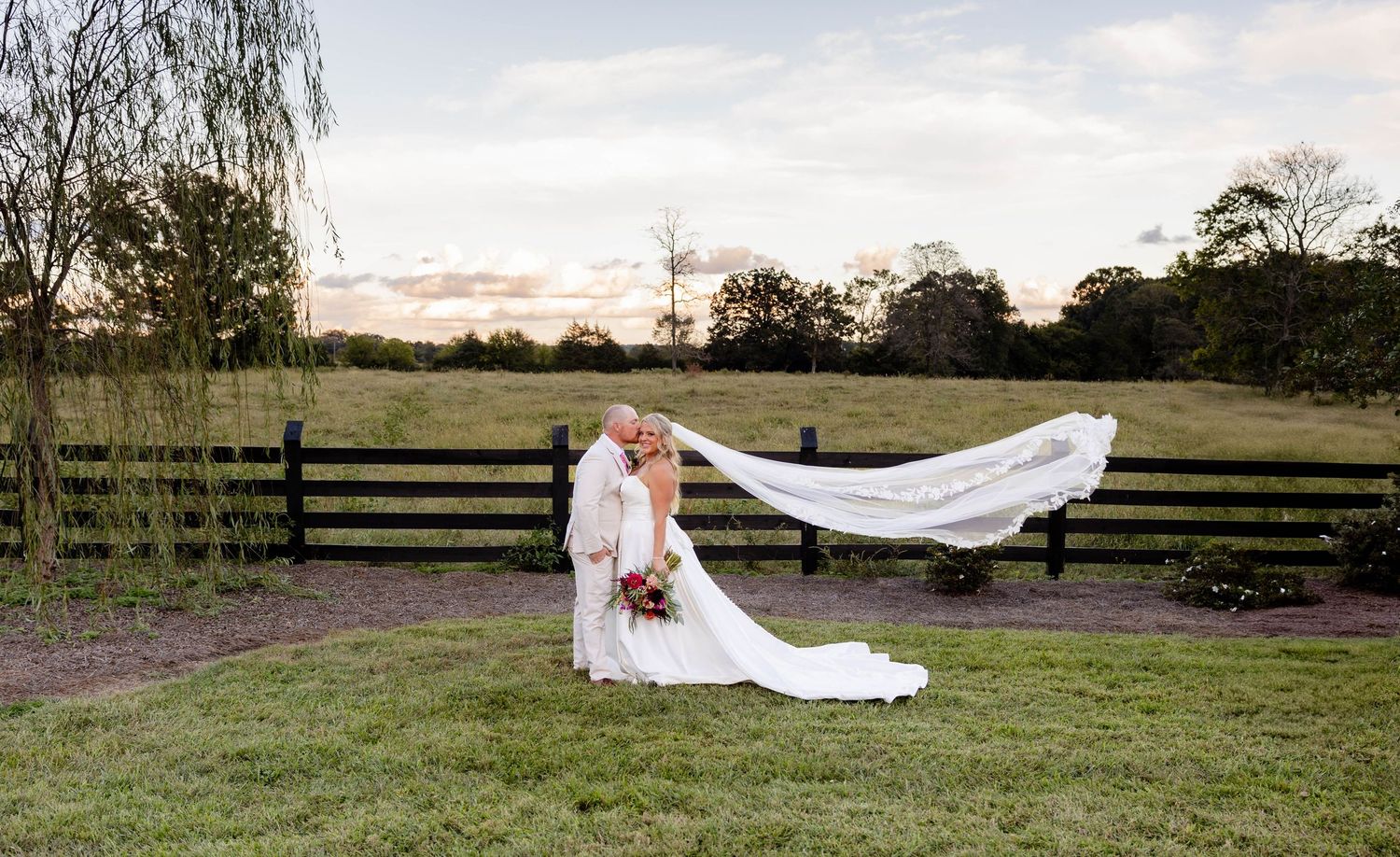 Wedding couple lying in grass with flowing veil spread out against rural fence backdrop.