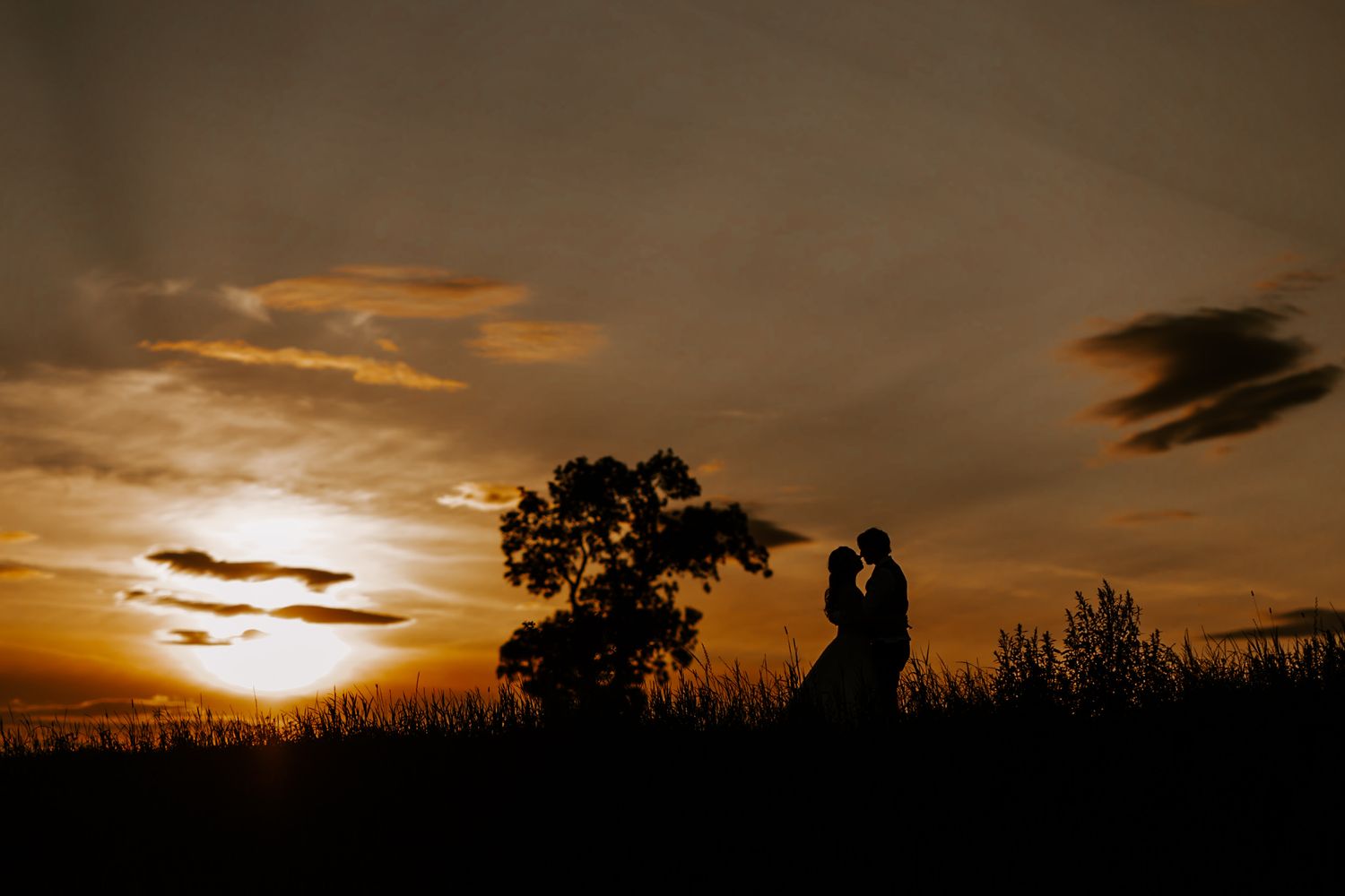 Silhouetted figures stand against a dramatic orange sunset sky with trees and clouds creating a romantic atmosphere.