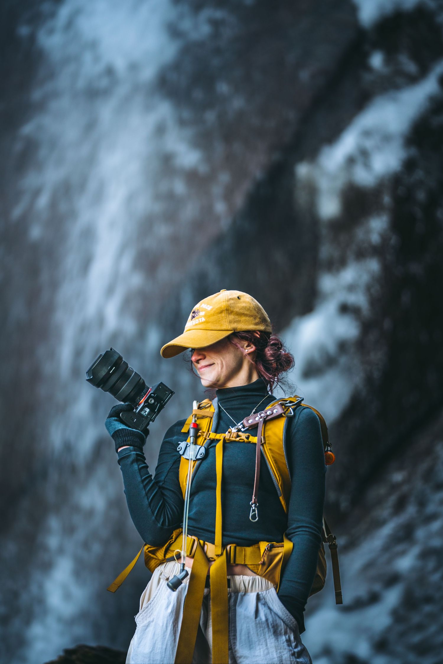 Female adventurer in yellow cap holding a camera while standing before a cascading waterfall.