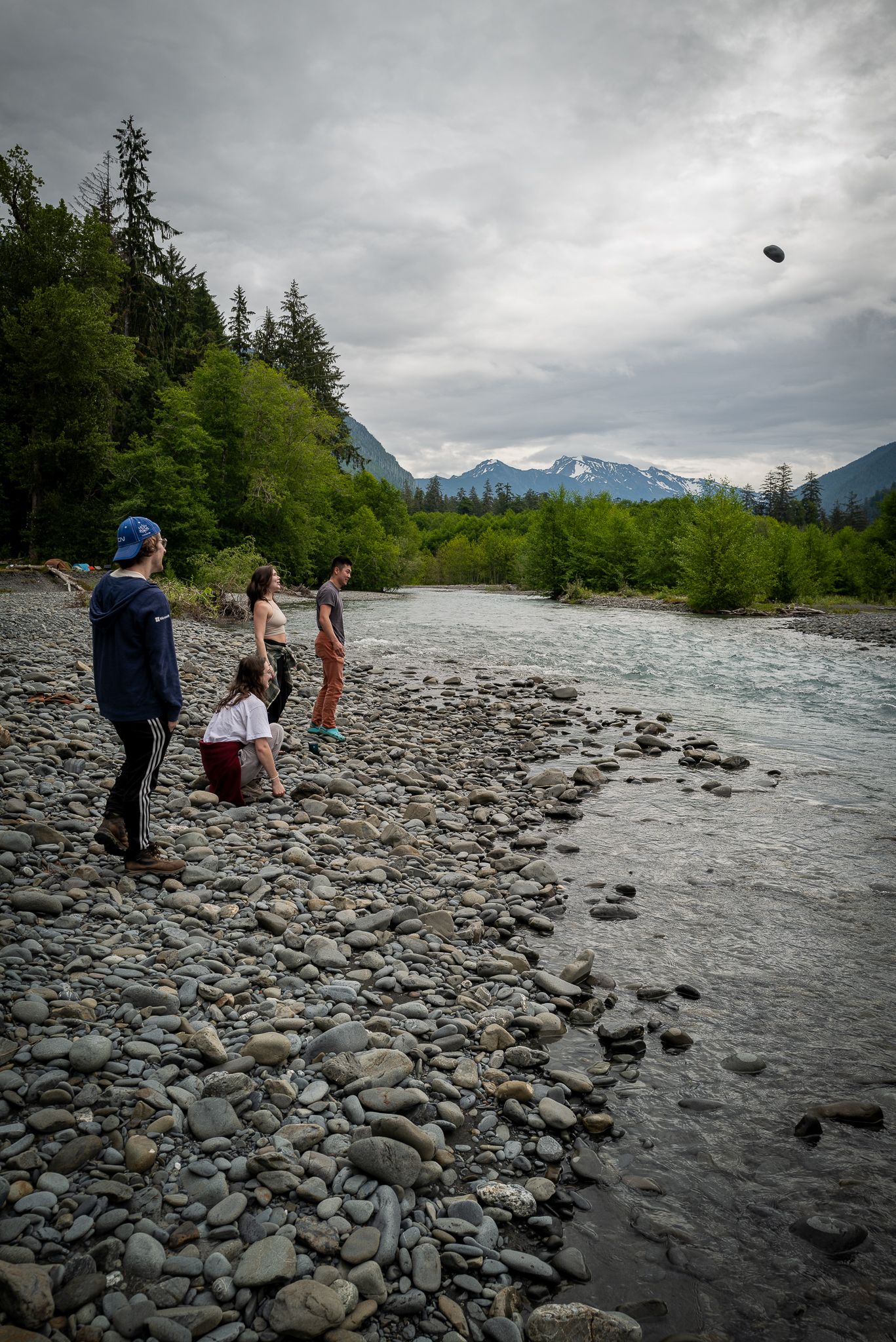 Group of people skipping stones on a rocky riverbank with mountains and evergreen trees in the background.