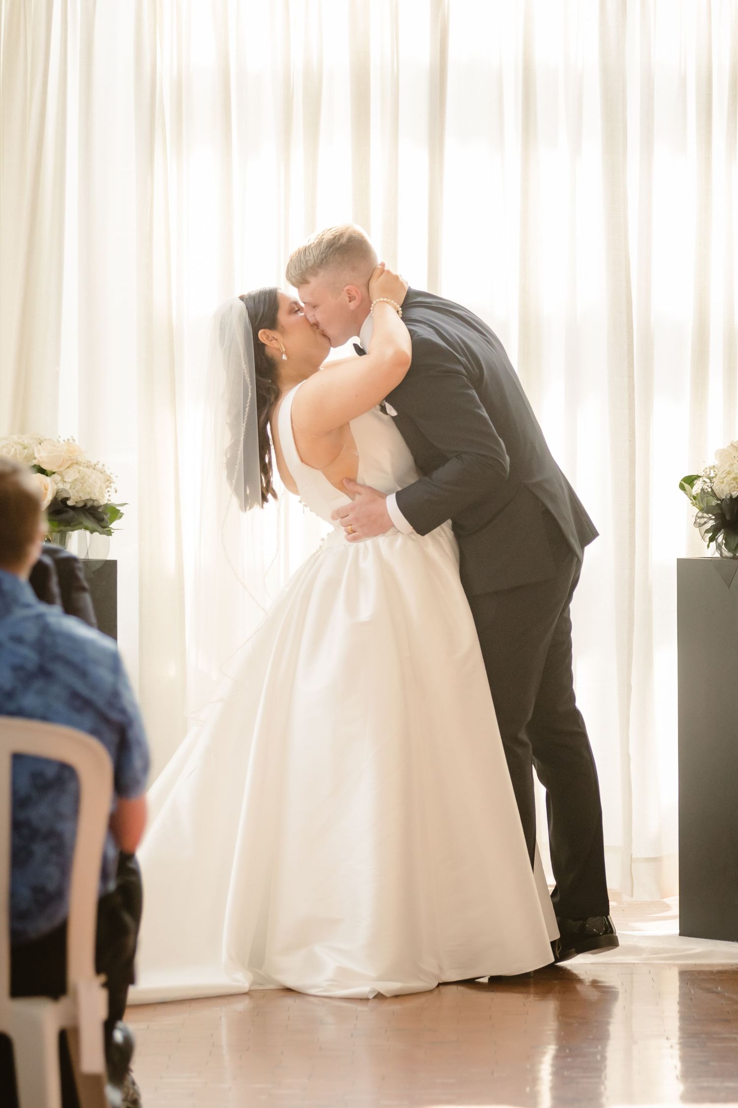 A couple shares their first kiss during an intimate wedding ceremony with white curtains and floral decorations.