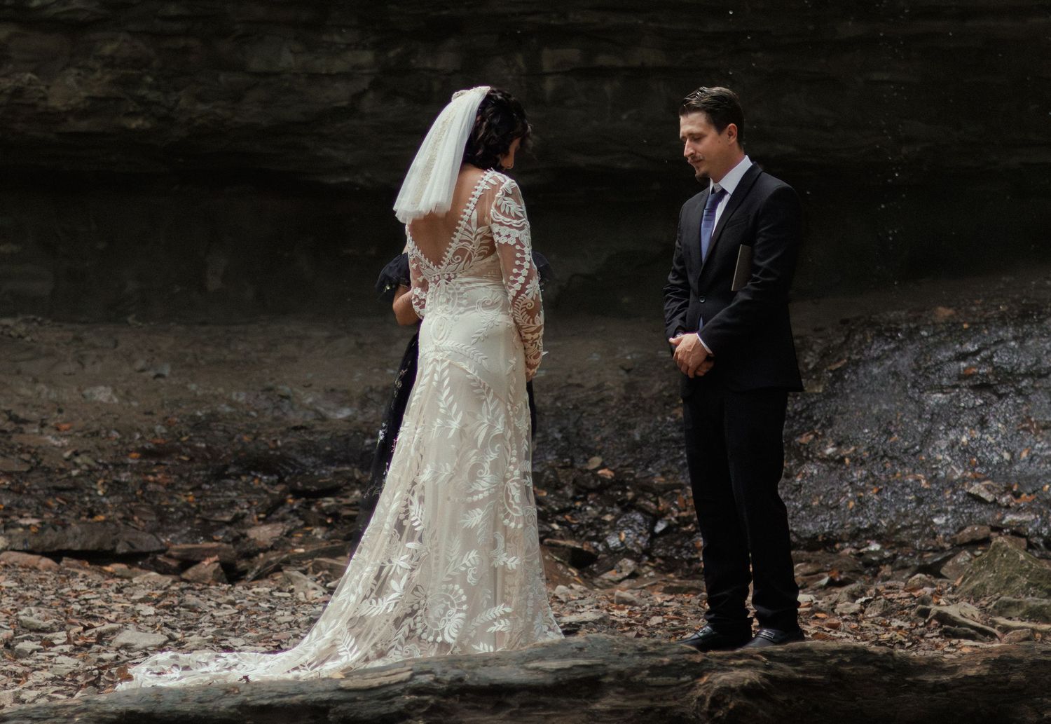 Bride in lace gown and veil faces groom in dark suit during intimate outdoor wedding ceremony near rocky cliffs.