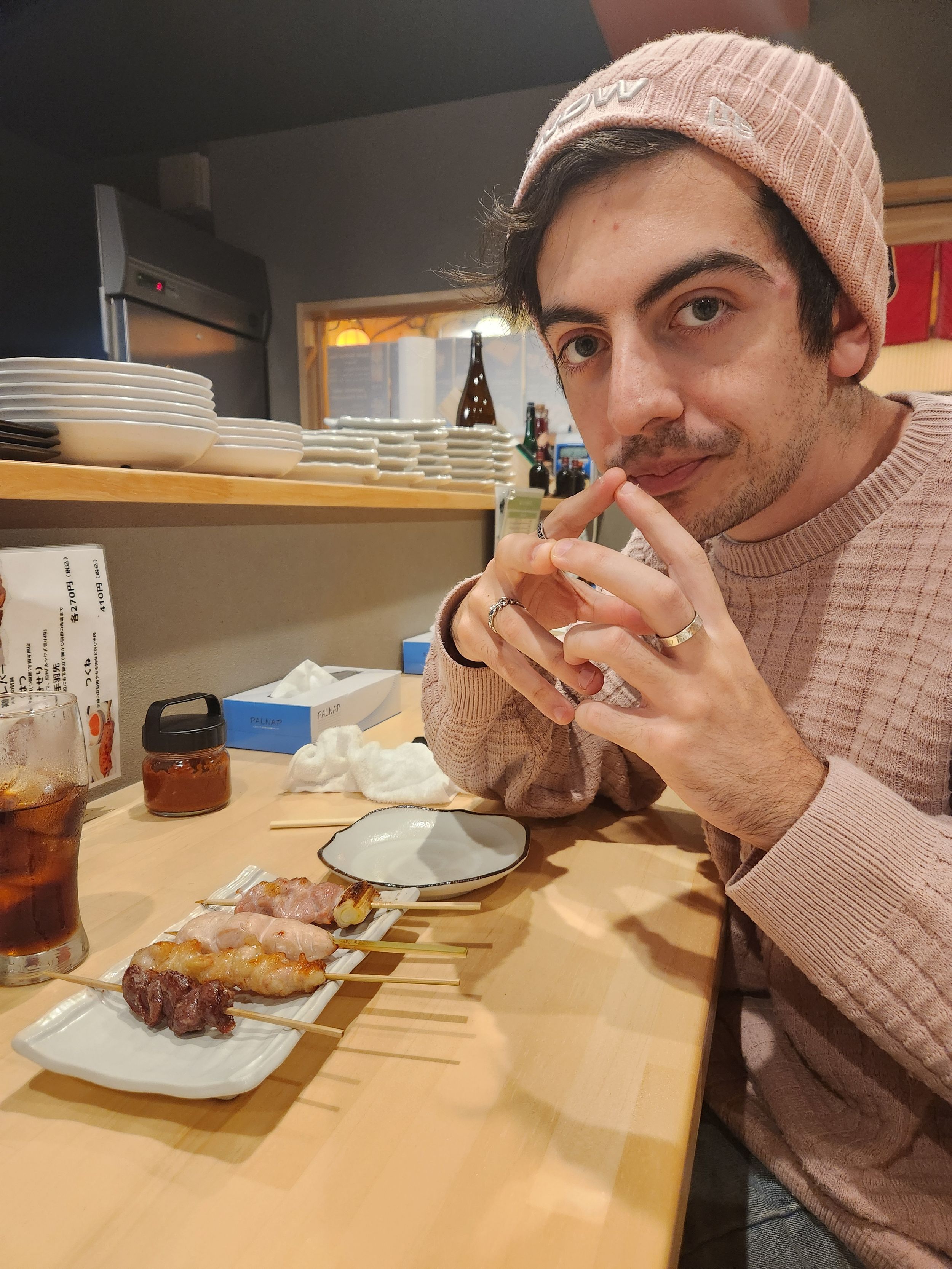 A person wearing a pink beanie eats a meal at a wooden counter in a dimly lit restaurant setting.