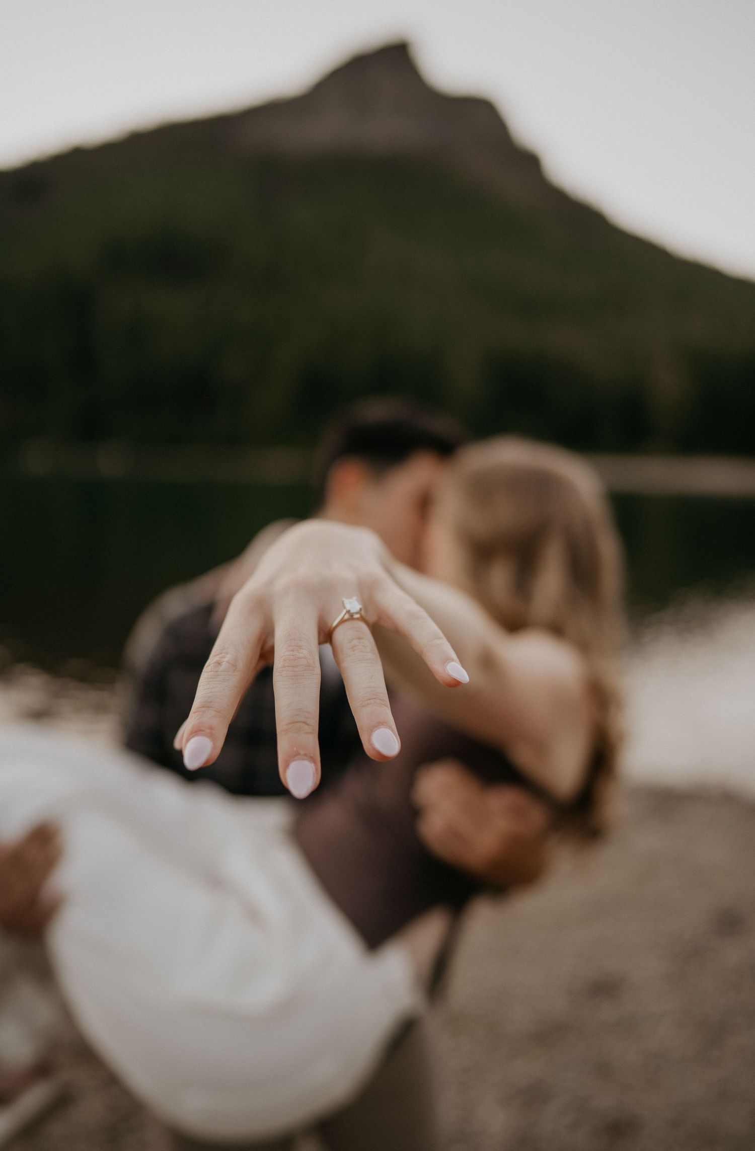 Newly engaged white couple flashing brand new engagement ring as they share a kiss on the cheek at Discovery Park WA