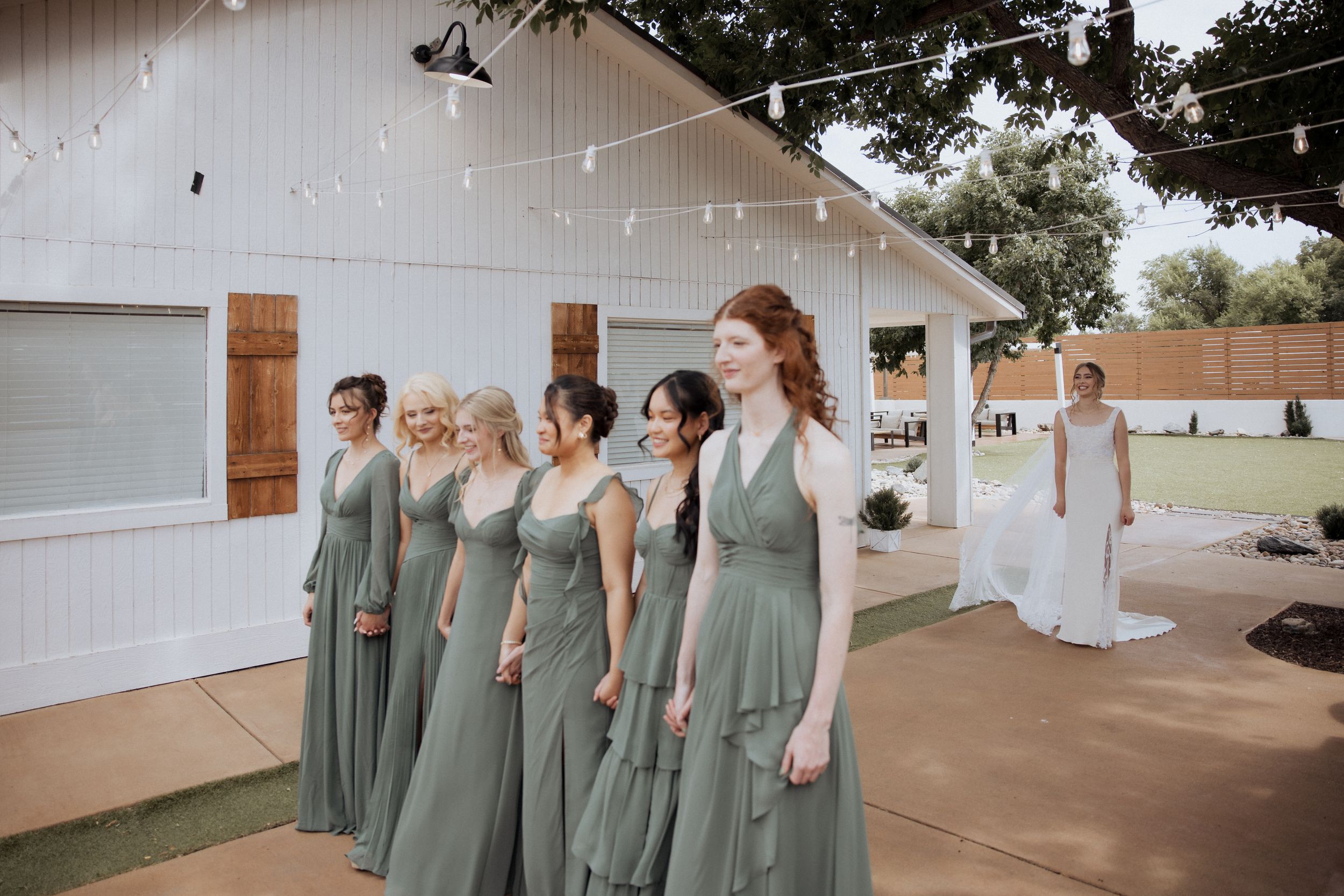 Wedding party in sage green bridesmaid dresses poses against a white building exterior.