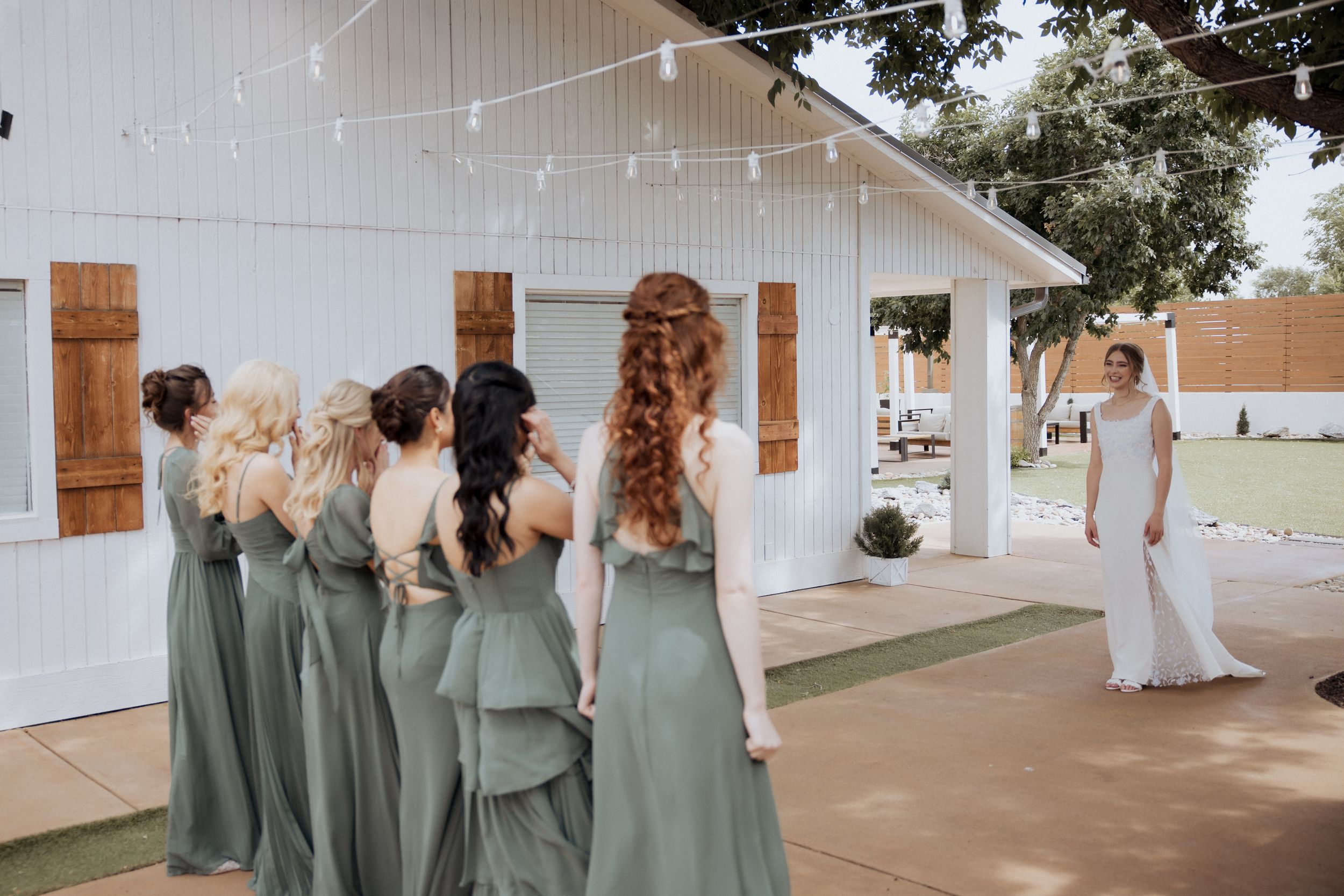 Bridesmaids in sage green dresses walk down a covered outdoor walkway at an elegant wedding venue.