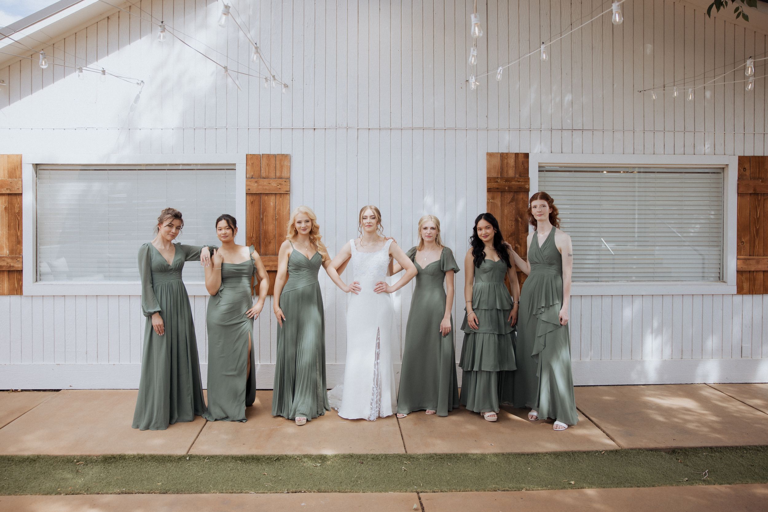 Bridal party wearing coordinated sage green gowns poses outdoors by modern white building.