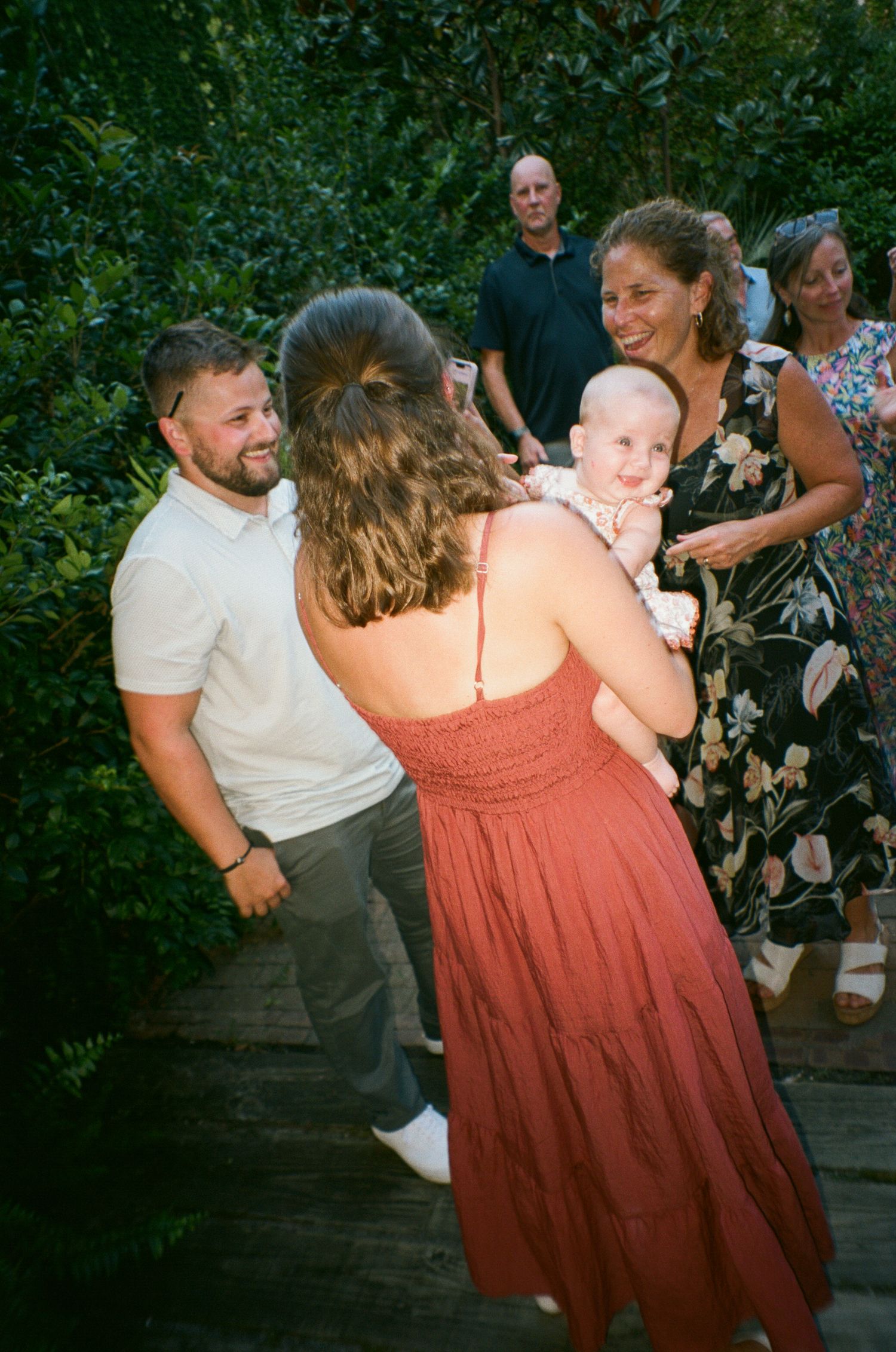 A group of people dancing and celebrating at an outdoor party with greenery in the background.