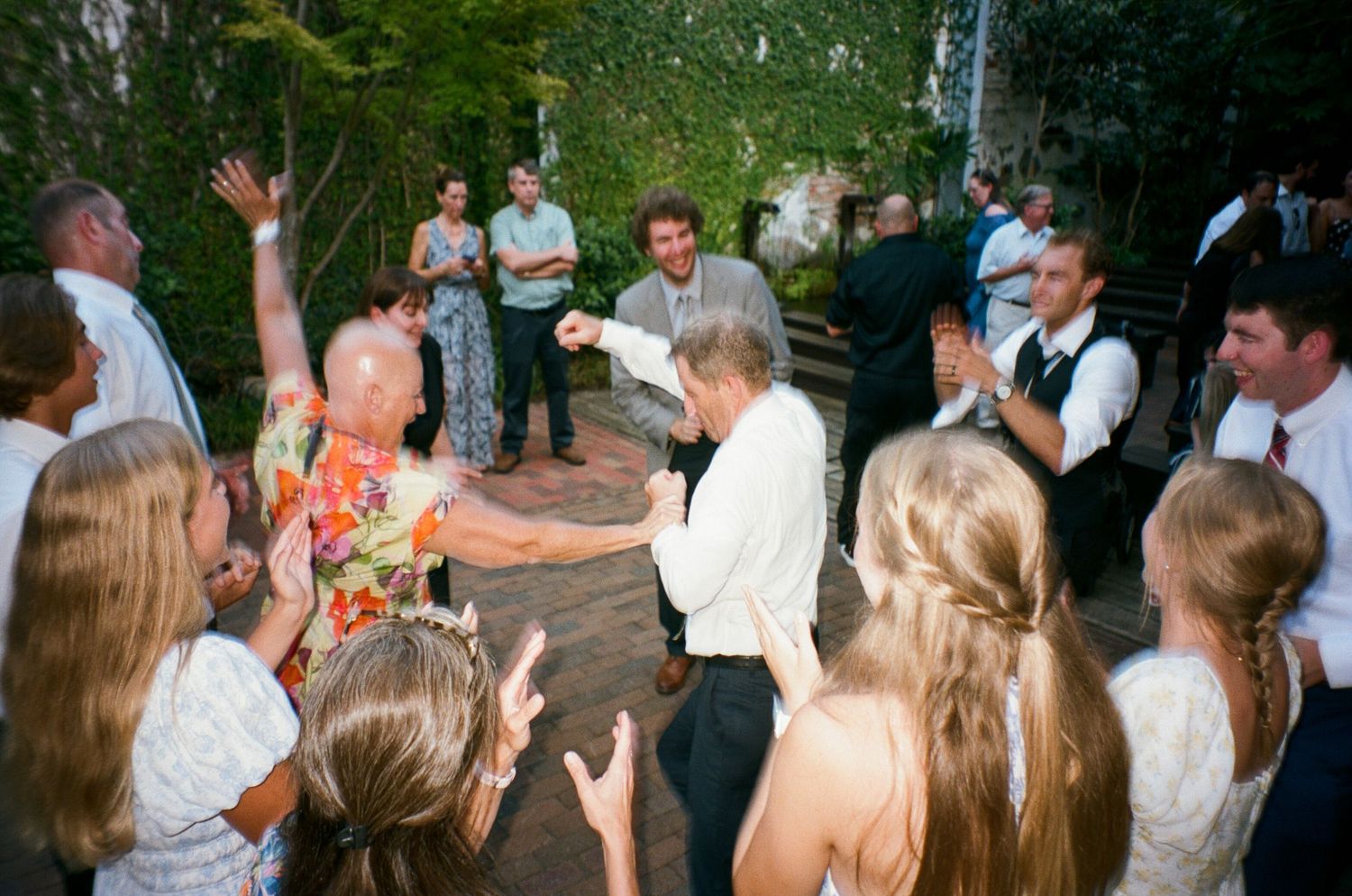 Wedding guests dance together during an outdoor reception celebration in a wooded setting.